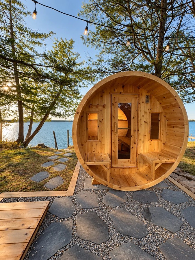 Cedar barrel sauna on a stone patio under string lights, steps from the water